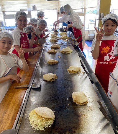 鉄板に並んだお好み焼と記念撮影する児童