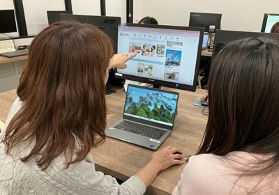 パソコンのモニターを指さし、隣の女性と話すFさん